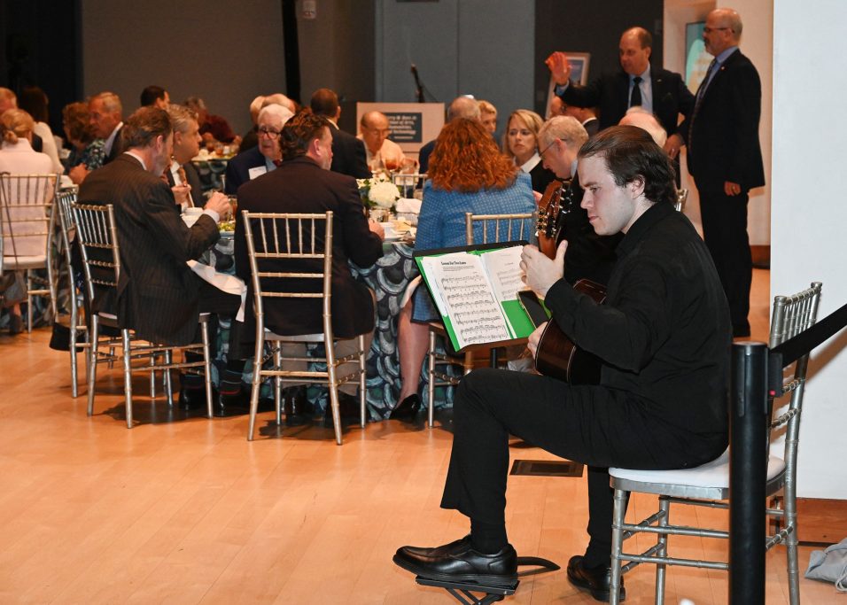 Person dressed in black playing a guitar. Group of people seated at a table.