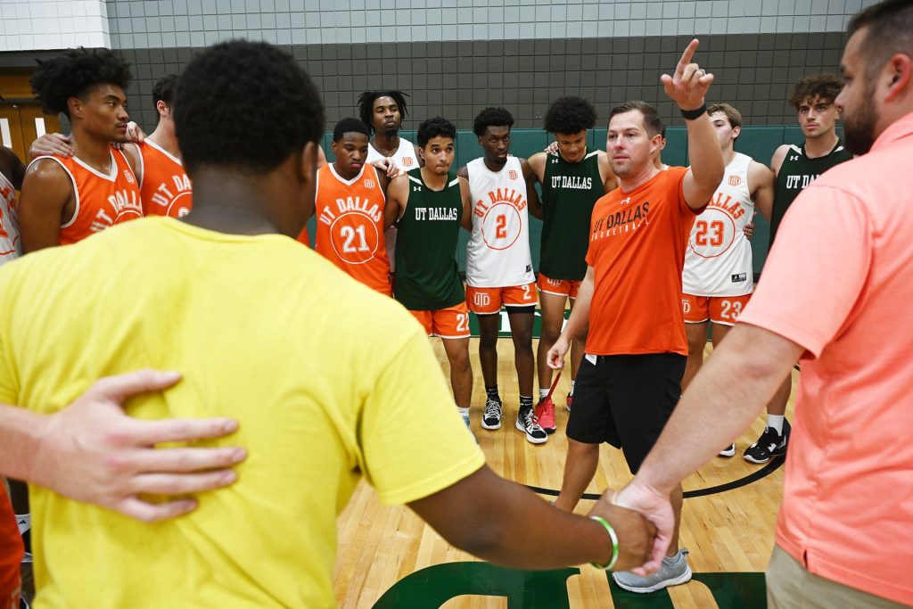 Group shot of UT Dallas basketball team. Coach Jared Flemming in orange shirt pointing.