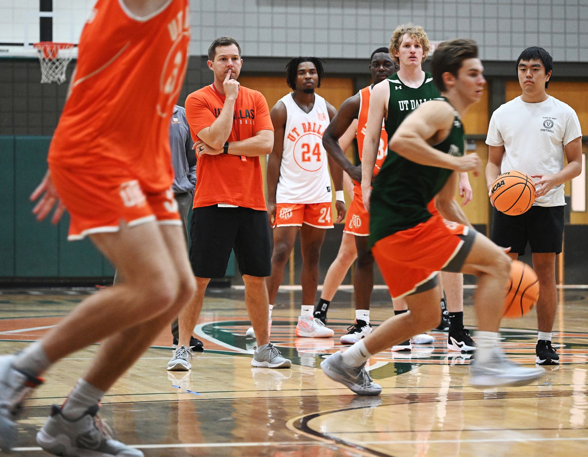 Action shot of UT Dallas basketball team. Coach Jared Flemming in orange shirt and dark green shorts holds finger to his lips as he observes.