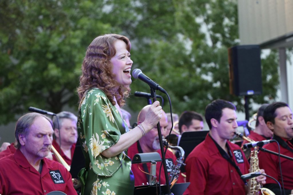 Woman singing into microphone with band playing in the background.