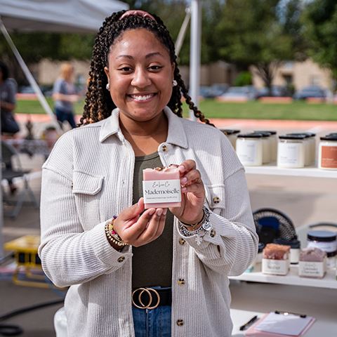 Homecoming event highlights alumni businesses. Woman in white coat holding up a homemade soap.