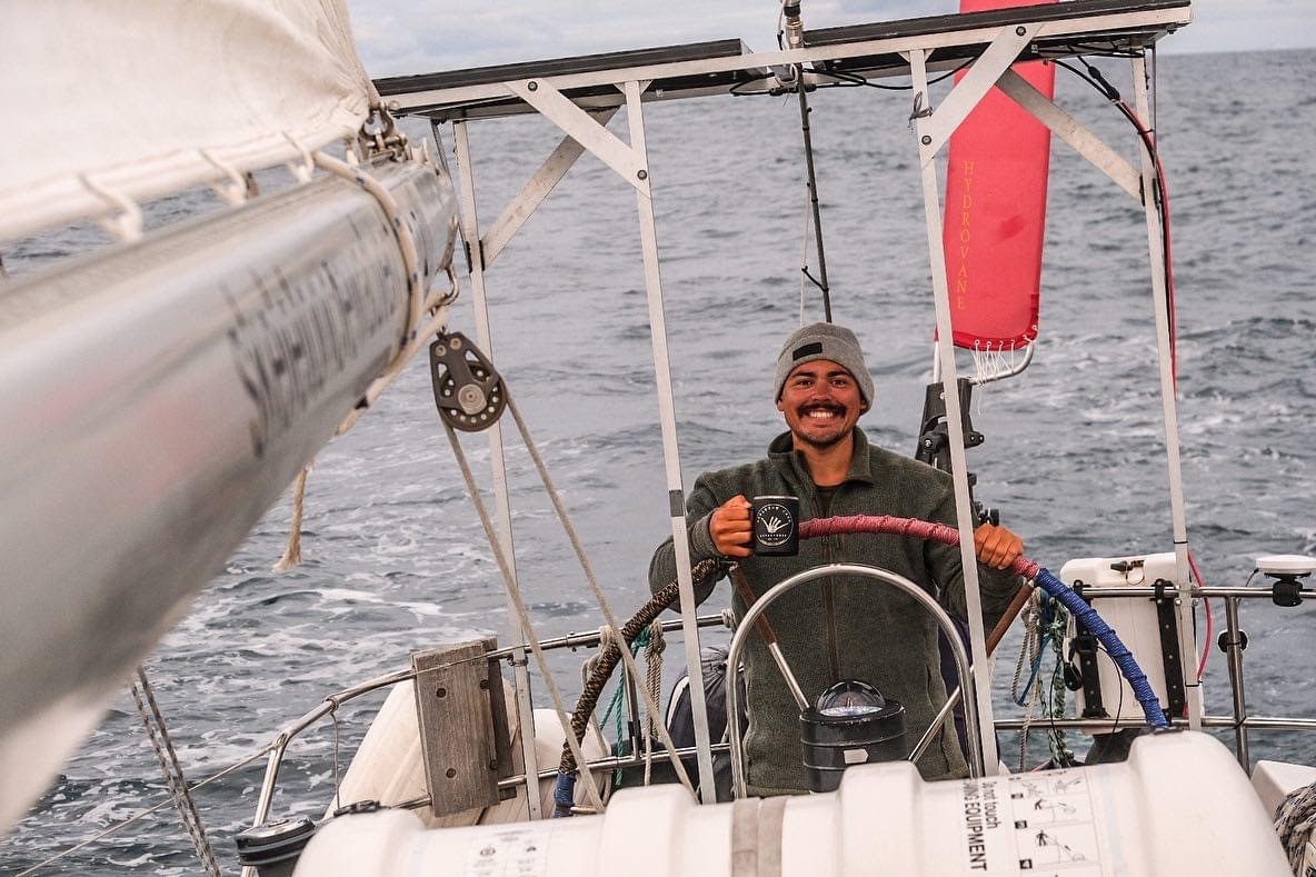 Ricky Gonzalez standing at the wheel of a sailboat while holding a cup of coffee.